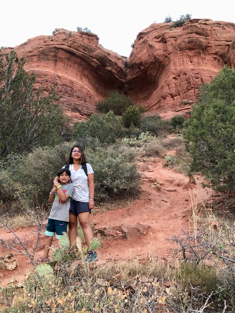 Boy and woman standing in from of a hill leading to a canyon cave of red rocks, with dry shrubs, bushes and desert landscape around the trail leading up to the cave.