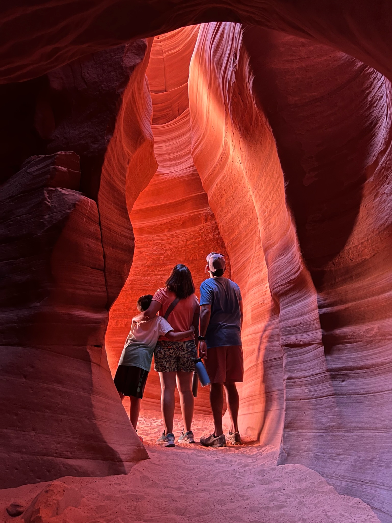 Boy, woman and man standing together with back towards camera looking up, insider a slot canyon with red sand and red canyon walls that look like painted strokes.