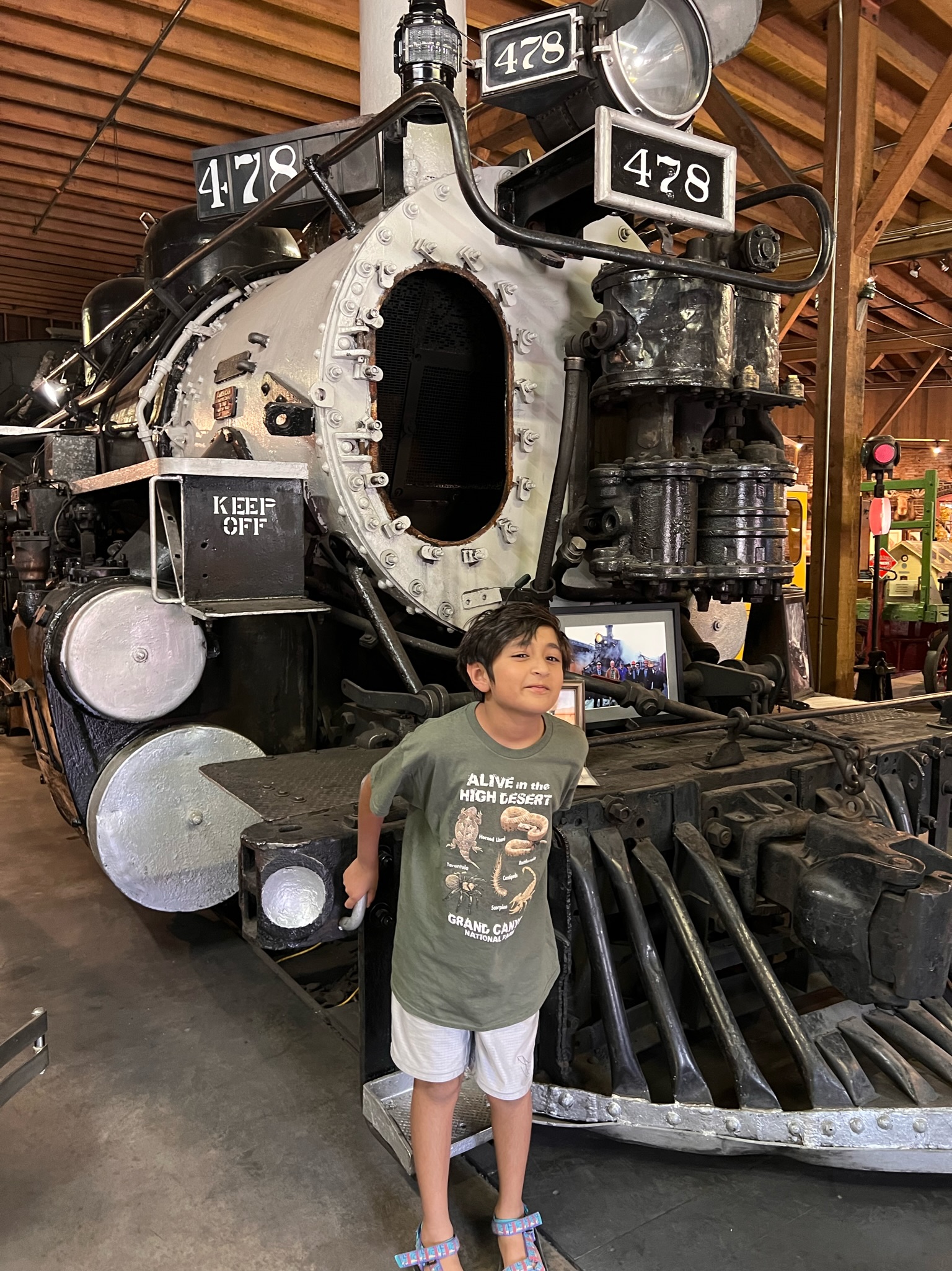 Boy standing in front of a vintage train engine of black and silver with an articulated engine and pipelines.