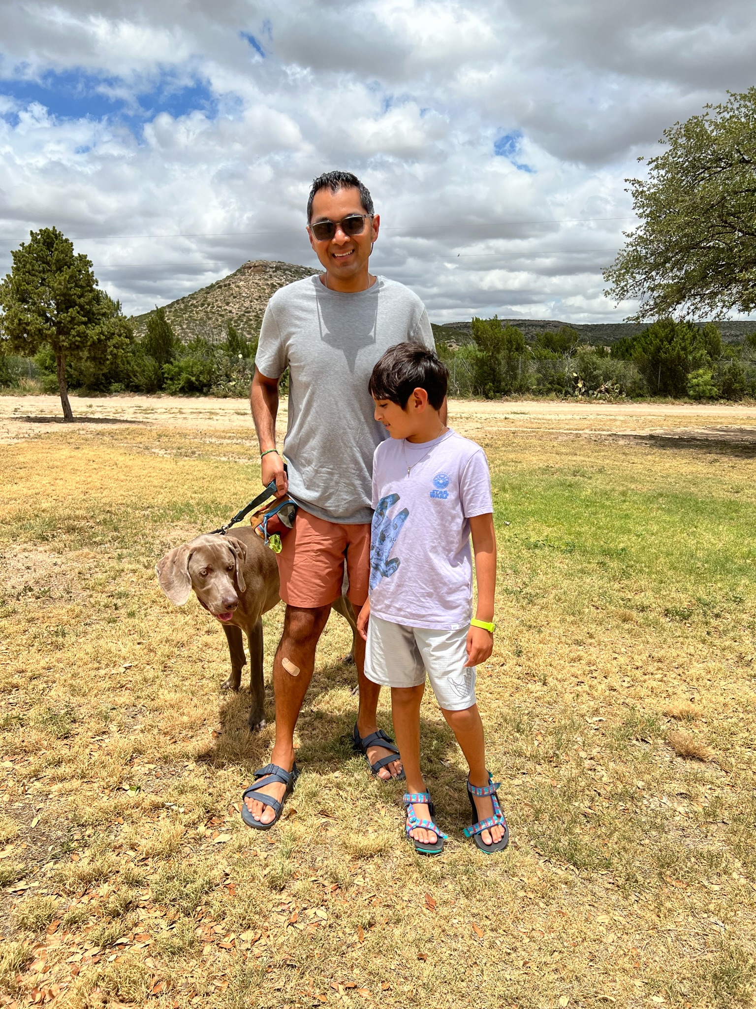 Dog, boy and man standing on dry grass field, with a hill in the distance. The geography looks shrubby with small trees in the background.