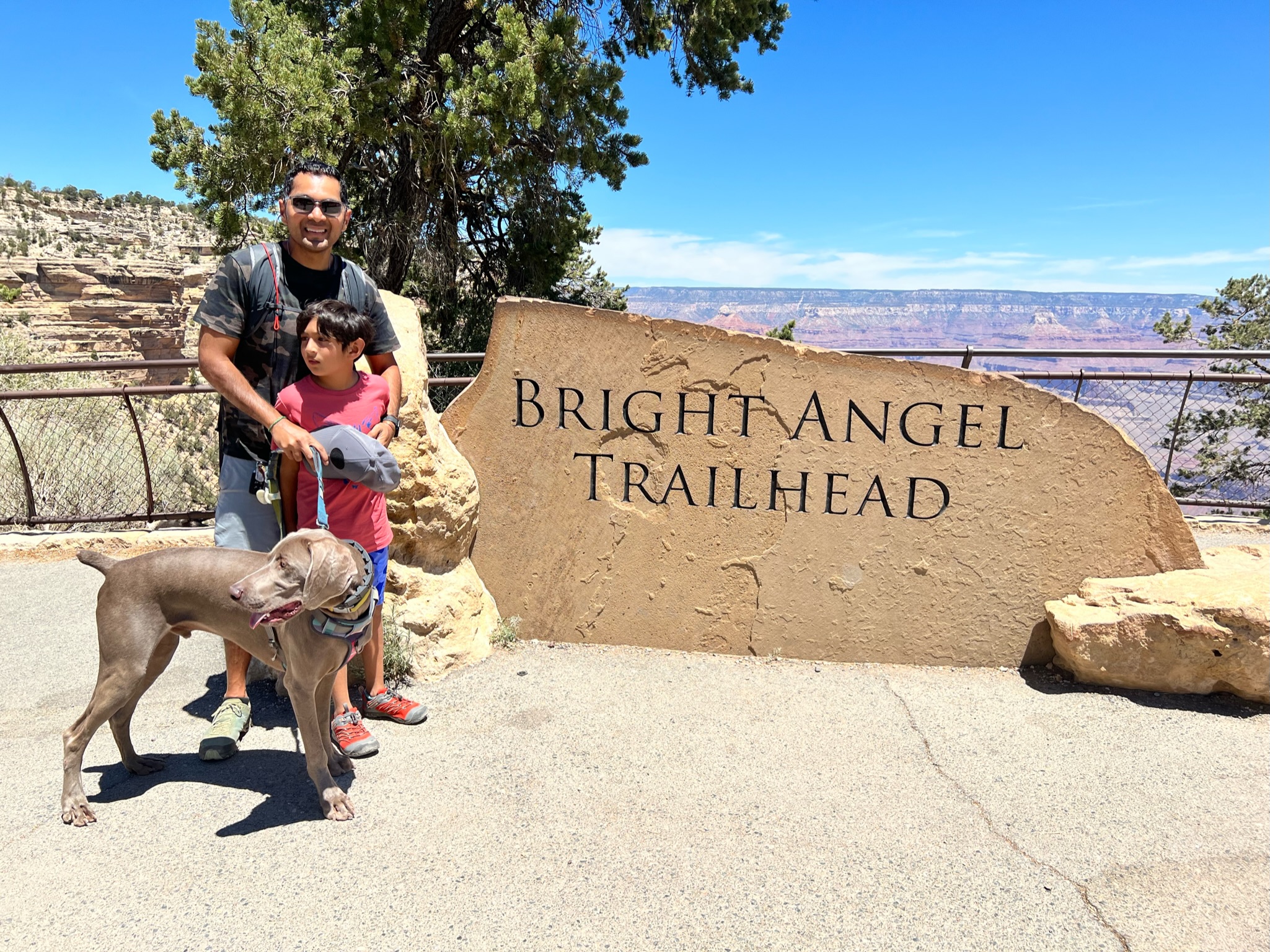 Man, boy and dog in front of a rock slab that says Bright Angel Trailhead, with Grand canyon views of red, orange and beige canyon walls in the background