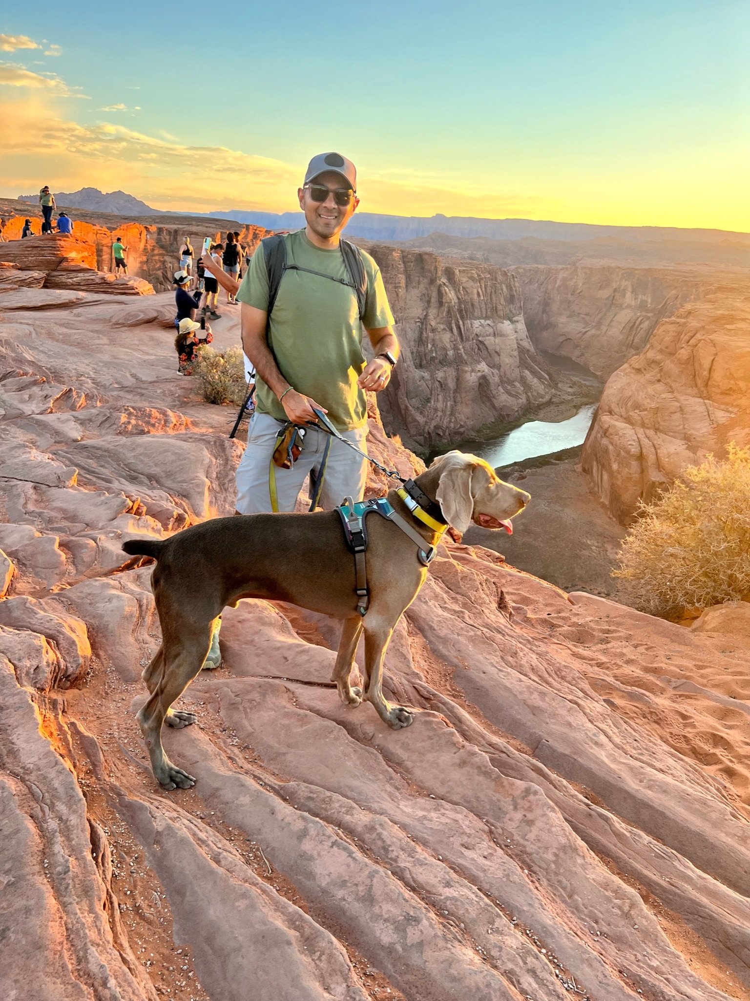 Man and dog standing on red canyon flowing slabs of rock, with horse shoe shaped snaking river with red canyon walls and desert landscape all around.