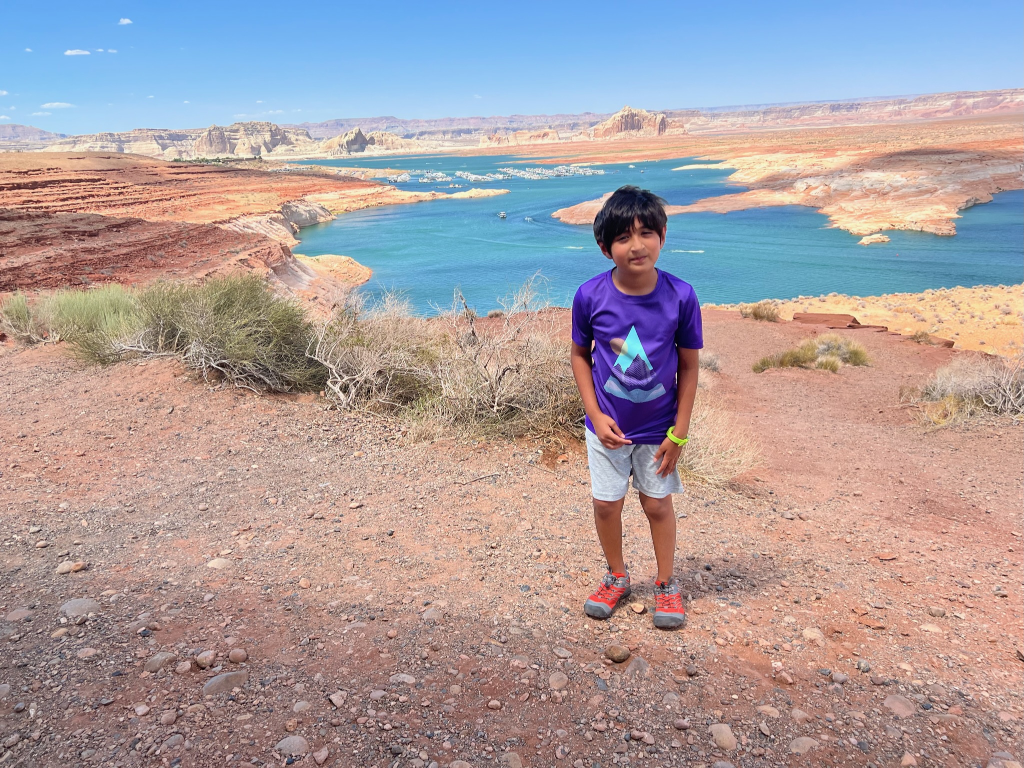 Boy standing on crush red gravel vista point with red canyons and all around and snaking blue lake in the middle