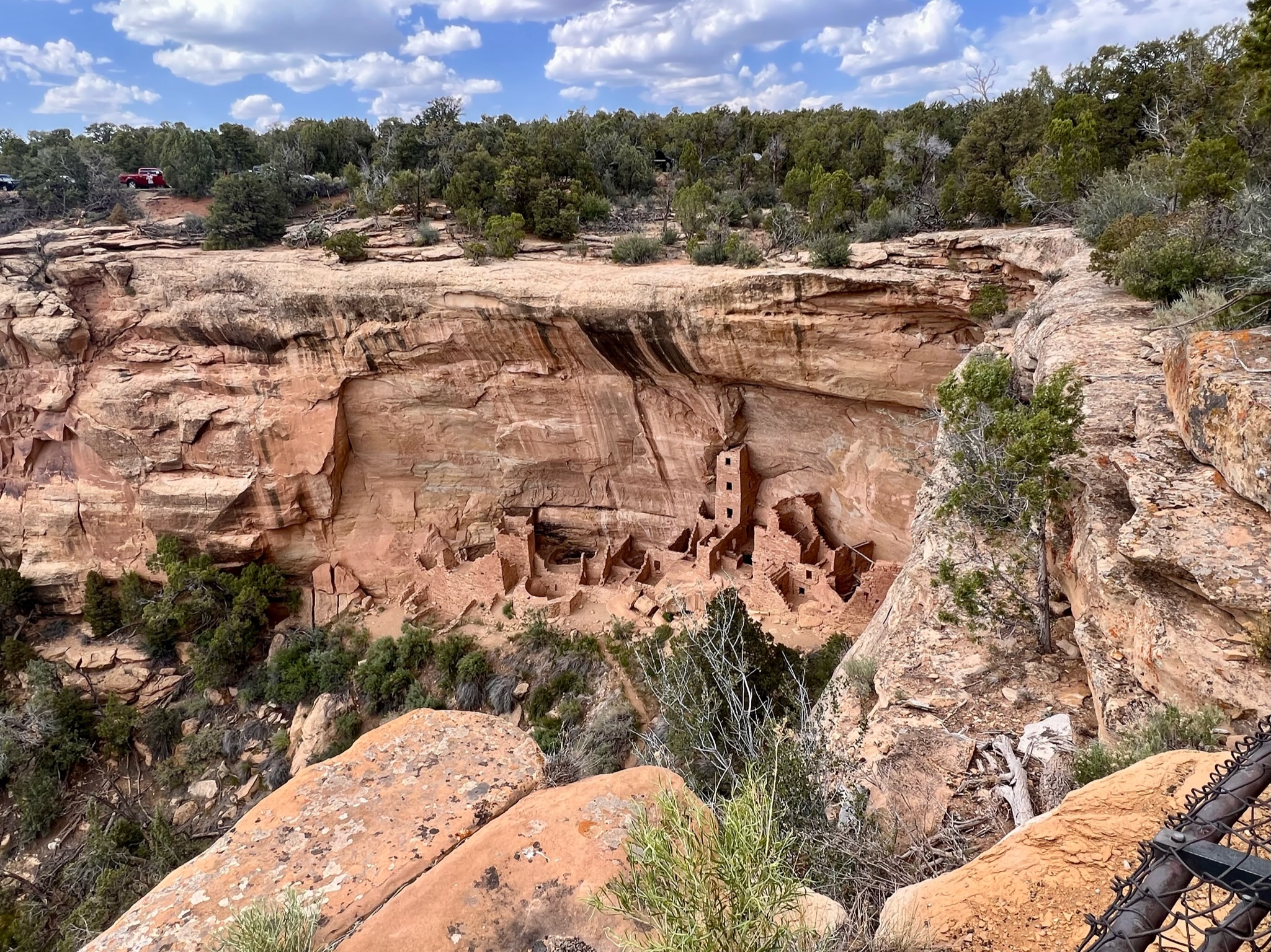 Red and beige canyon walls with top of green desert plants, shrubs and trees. The wall side has the remains of living spaces and tower of a civilization that used to live hundreds of years ago.