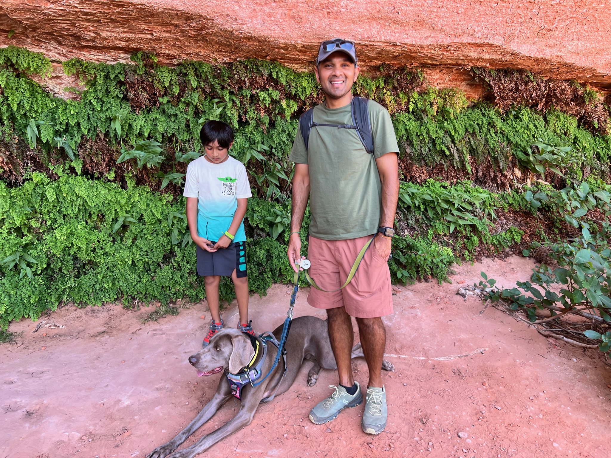 Man, boy and dog in front of red canyon wall and slabs of rock, with the wall covered with green ferns in a cove looking place.