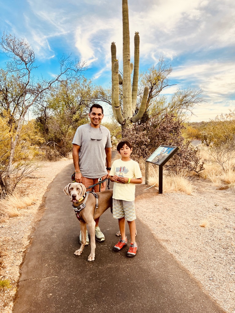 Dog, boy and man standing on a paved walk path with a large Saguaro and other desert trees and shrubs in the background.