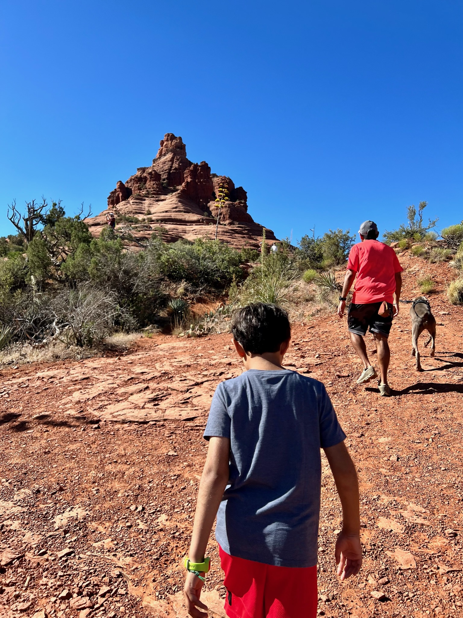 Man, dog and boy climbing up red rock hill with crushed gravel beneath and bell shaped rugged rock formation at the top of the hill.