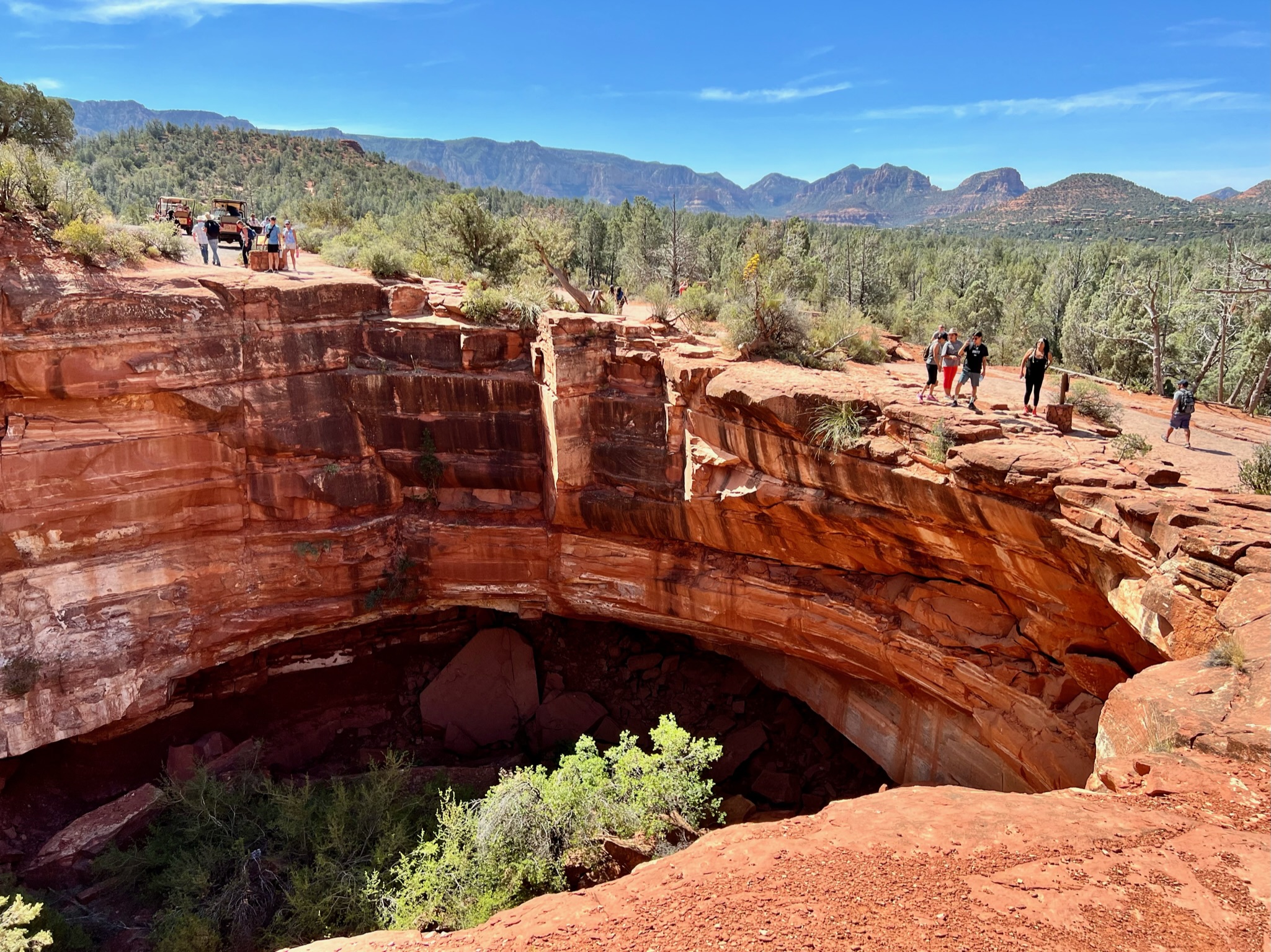 Huge red rock formations with a sinkhole in the middle. Lots of desert vegetation growing in the rocky hilly landscape.