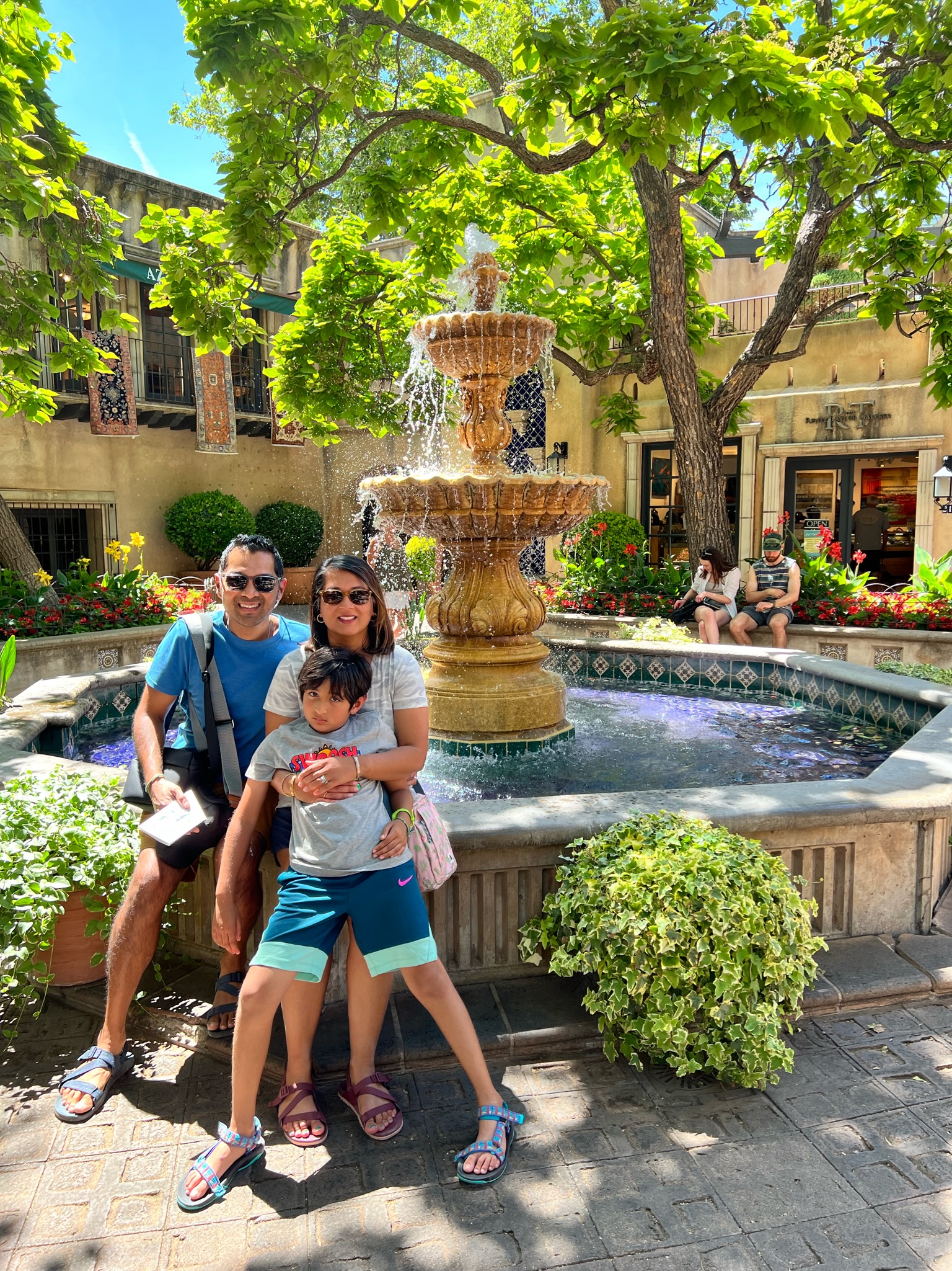 Man, woman and boy sitting in front of a water fountain, in the courtyard of a commerical shopping marketplace.