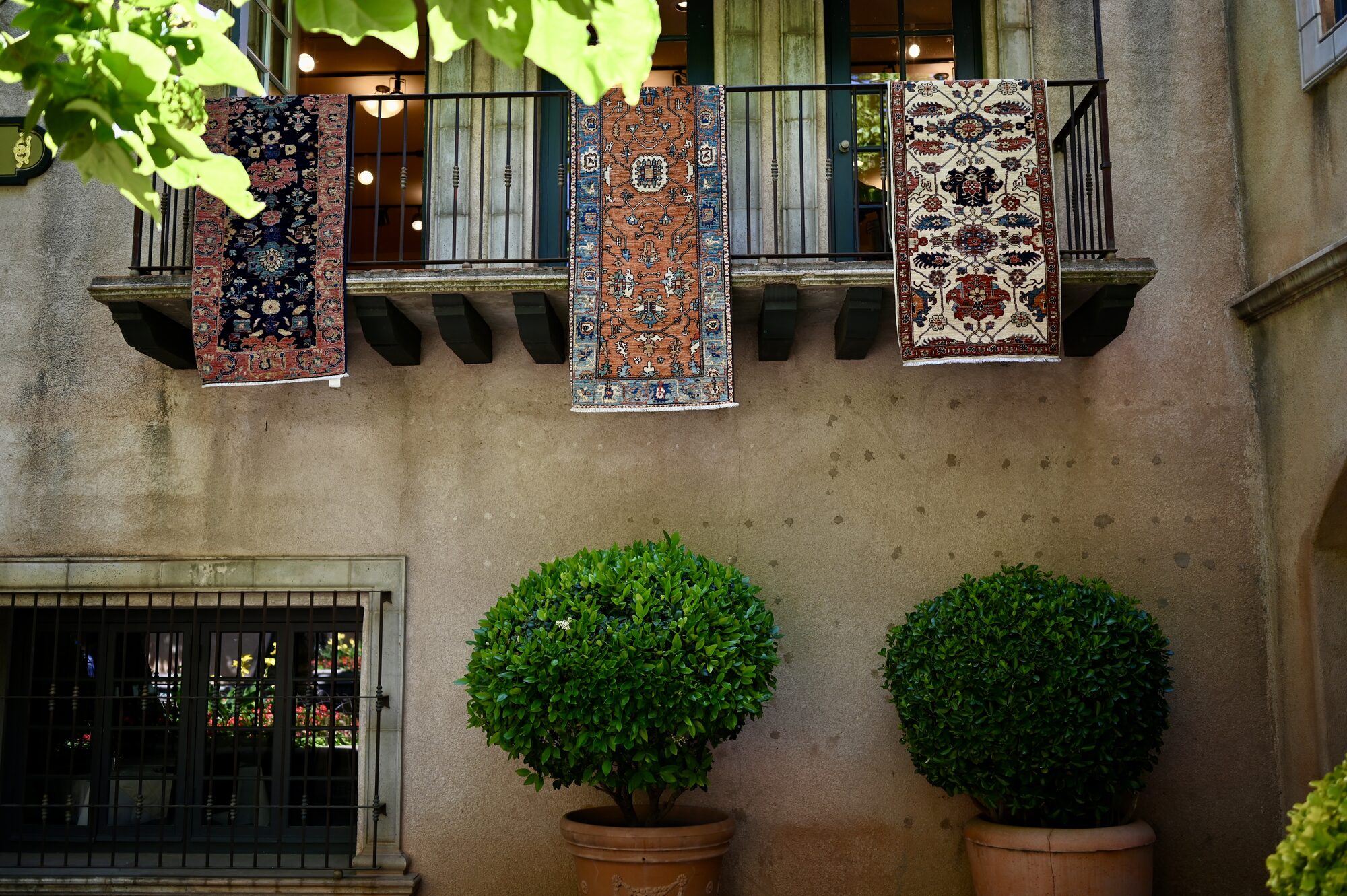 Patterned rugs hanging from a balcony over potted shrubs at Tlaquepaque