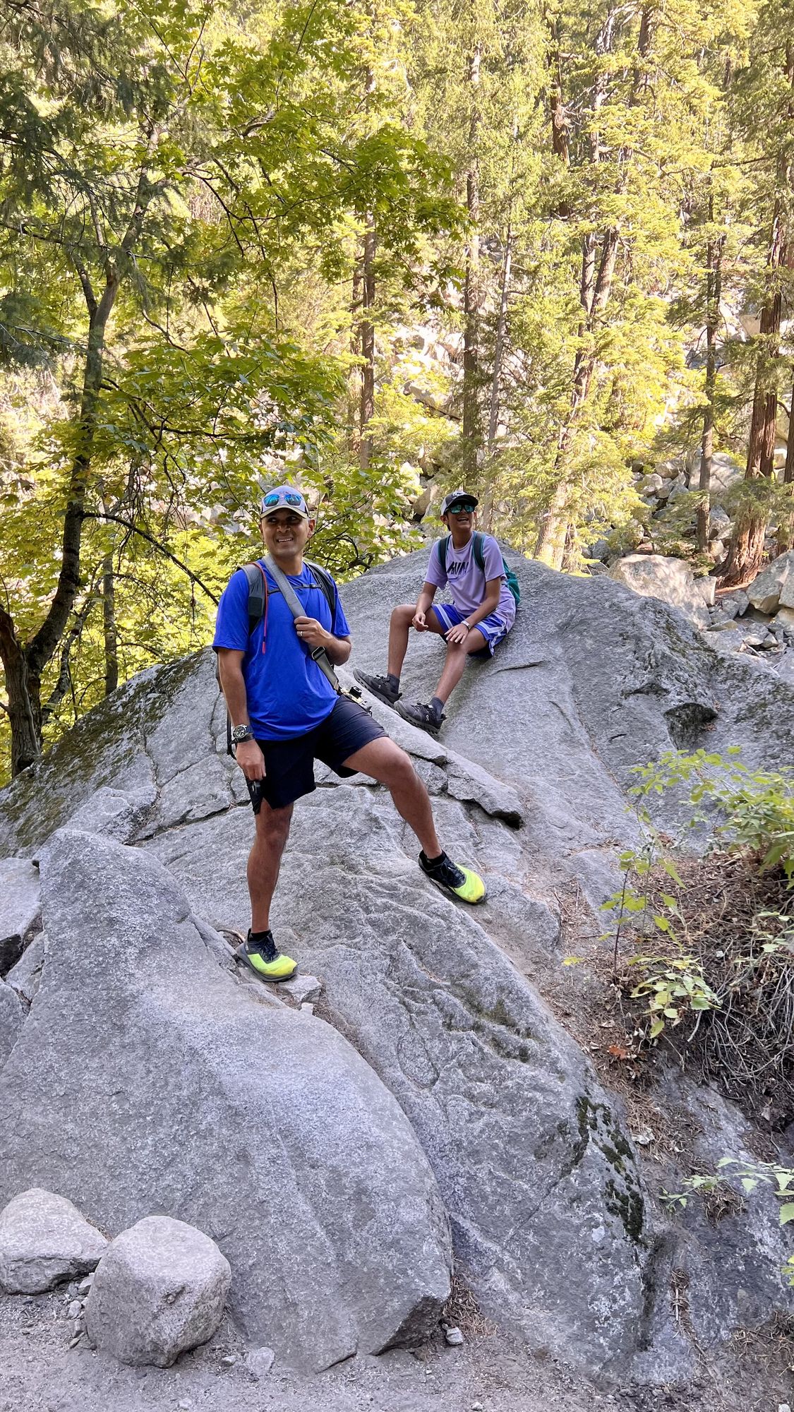 Our son and his dad resting on a large granite boulder beside the Mist Trail, surrounded by trees.
