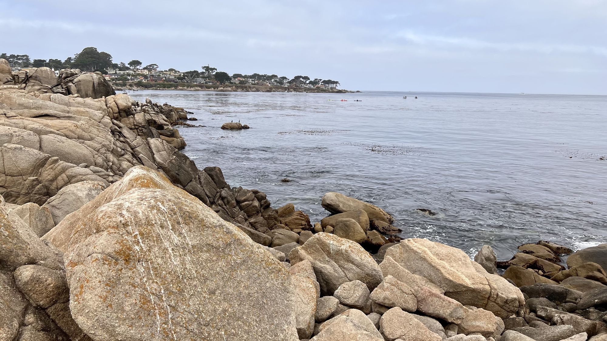 Large granite boulders along the shore, with calm grey water and houses on the far point.