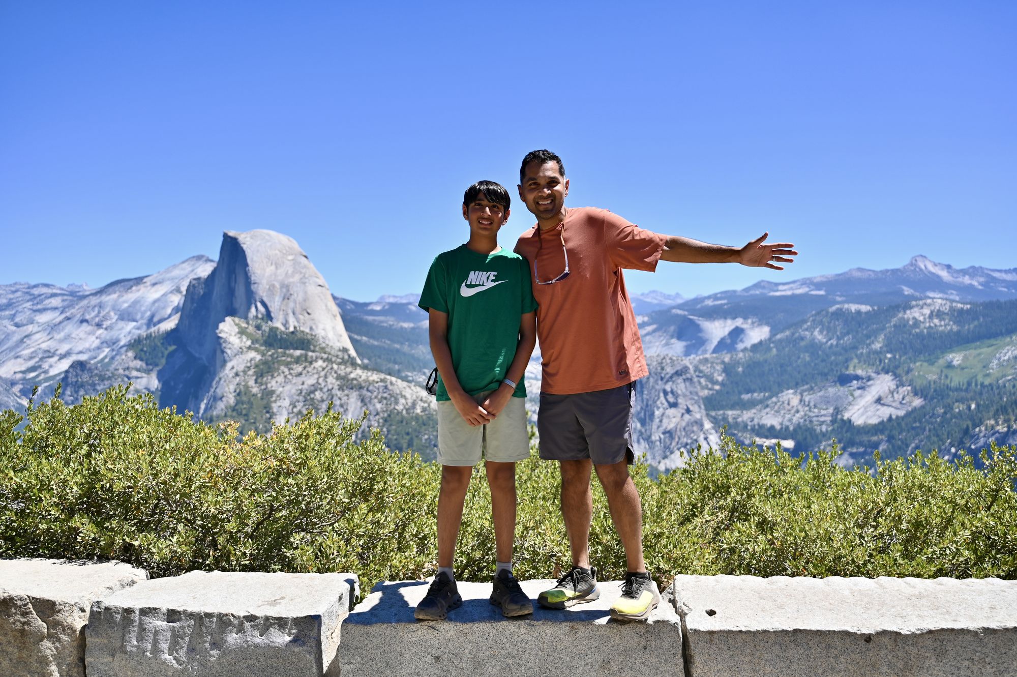 Our son and his dad standing on a stone wall at Glacier Point, with Half Dome behind them.