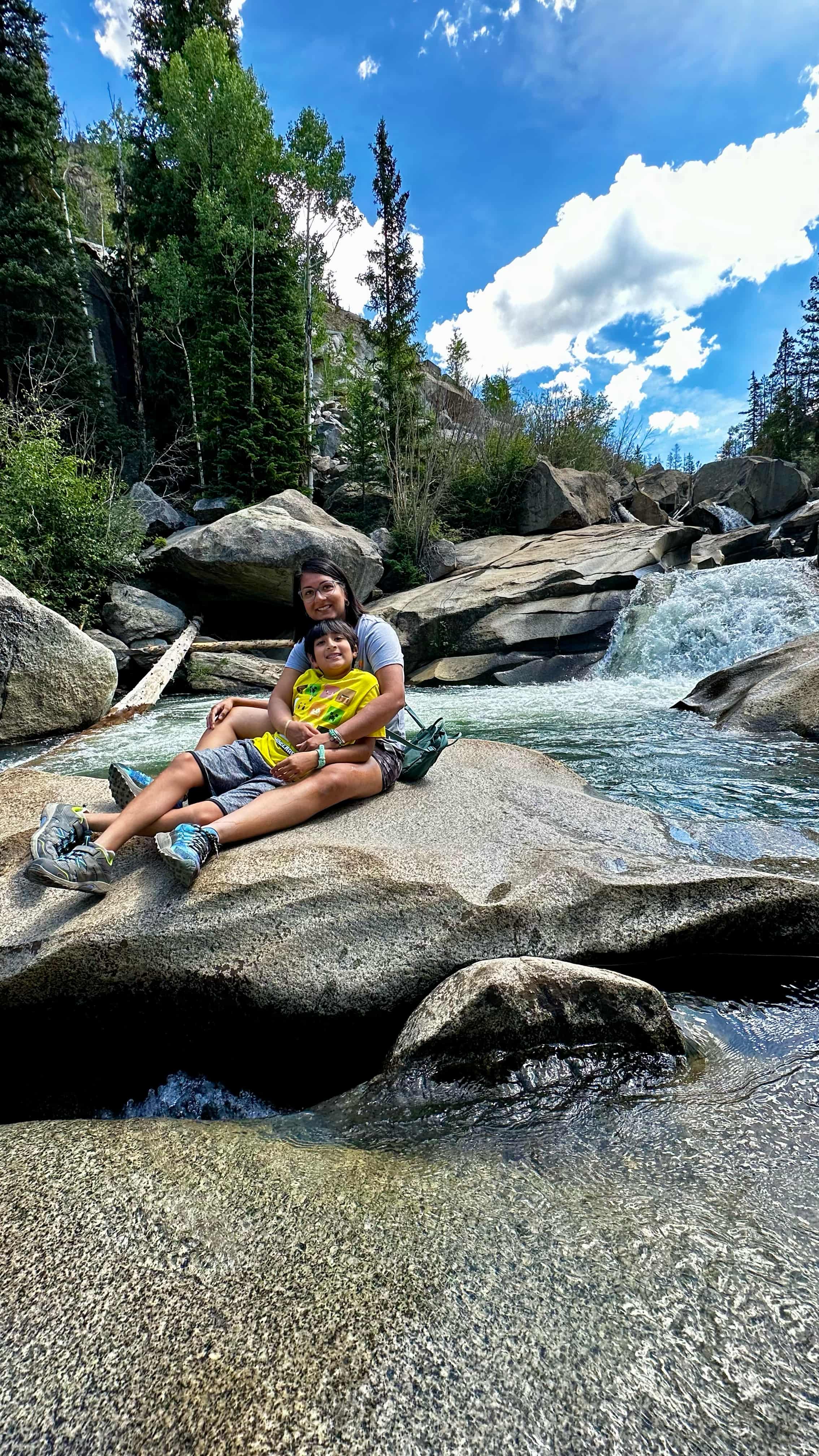 Mom and son sitting on the rocks by a river with waterfall, mountain and trees in the background