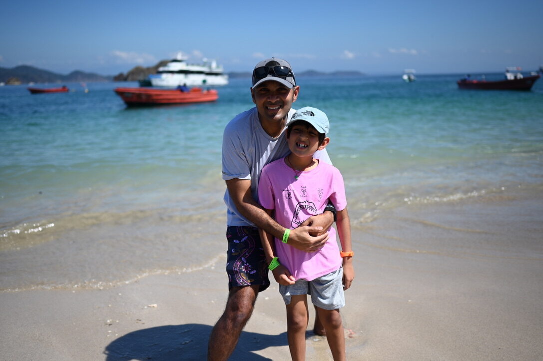 Man and boy hugging on the white sand beach, with boats in the turquoise water behind