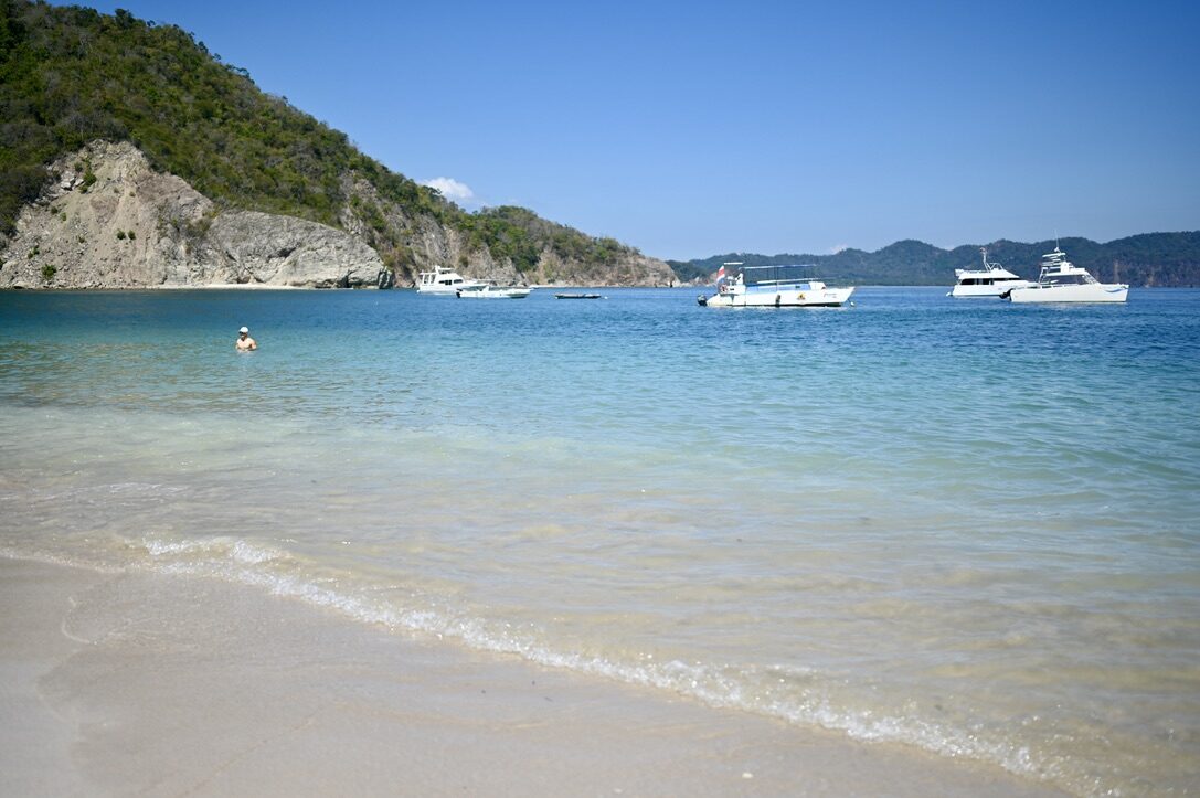 Clear turquoise water lapping the white sand, with boats anchored off the island