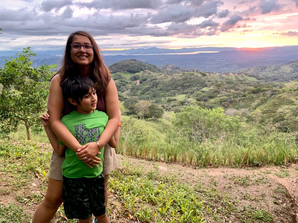 Woman hugging a boy on a hillside at sunset, with green hills rolling to the coast