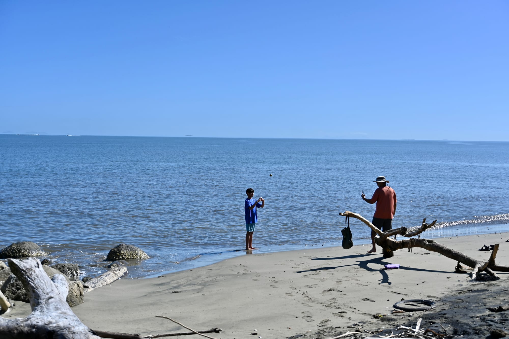 A narrow strip of grey sand scattered with driftwood and rocks, where our son and his dad stand at the water’s edge taking photos of the flat, calm sea.