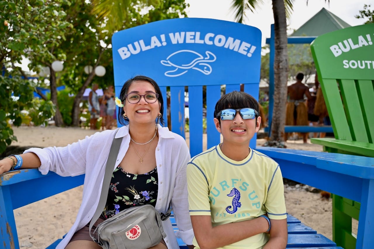 Our son and I sitting on a giant blue wooden chair painted with a turtle and “Bula! Welcome!”, with a green one beside it and dancers in grass skirts behind.