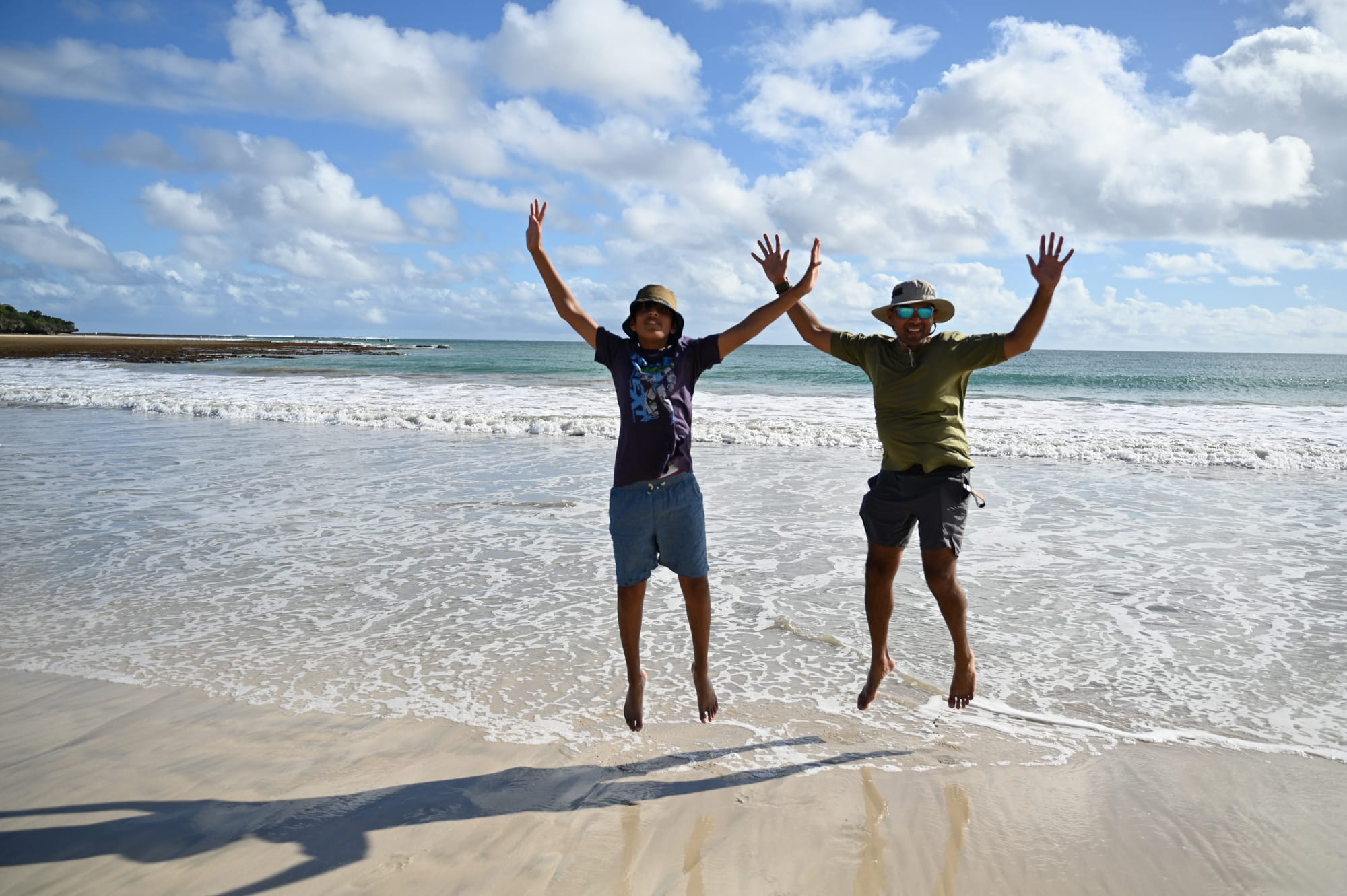 Our son and his dad leaping in the air with arms raised on wet sand, as white surf rolls in behind them.