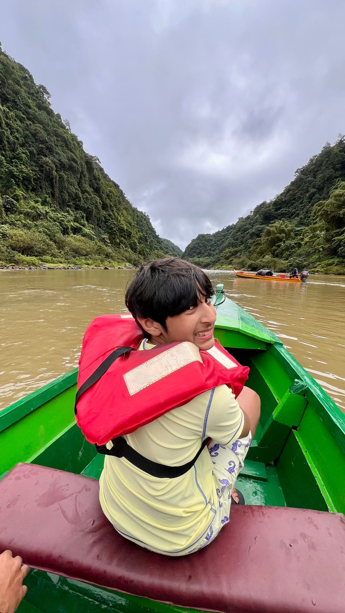 Our son in a red life jacket looking back from the bow of a green wooden longboat, on a wide brown river between steep forested hillsides under a grey sky.