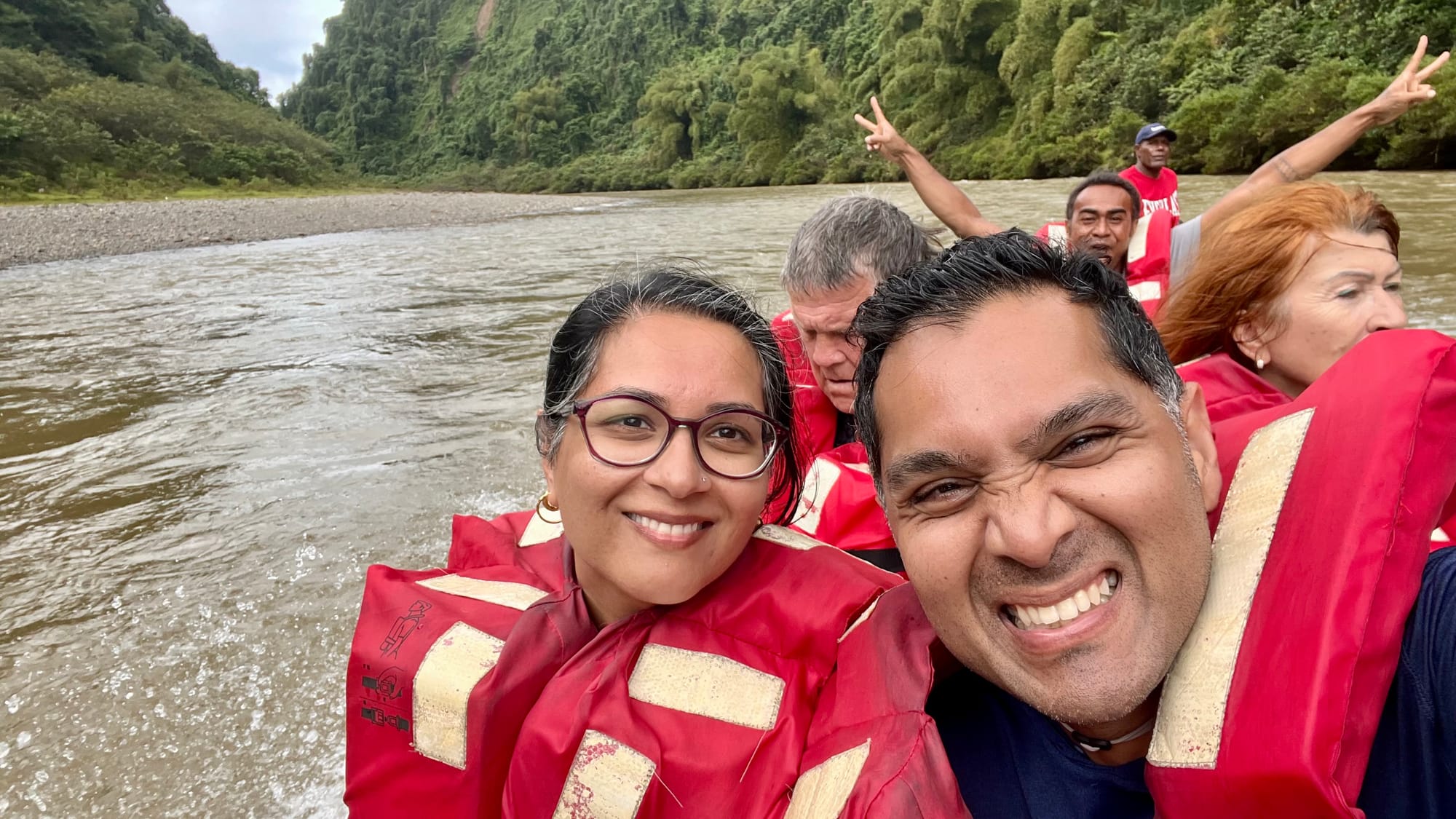A grinning selfie of me and my husband in red life jackets on the longboat, with other passengers waving behind us and the river and forest beyond.