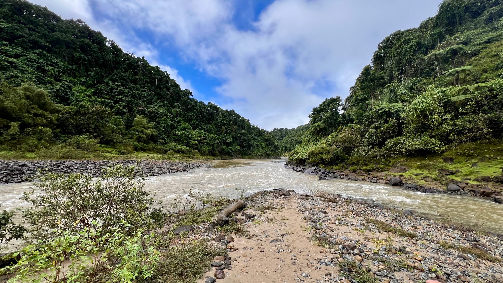 A rocky, gravel spit at a bend in the fast-flowing river, with dense green hills rising on both sides.