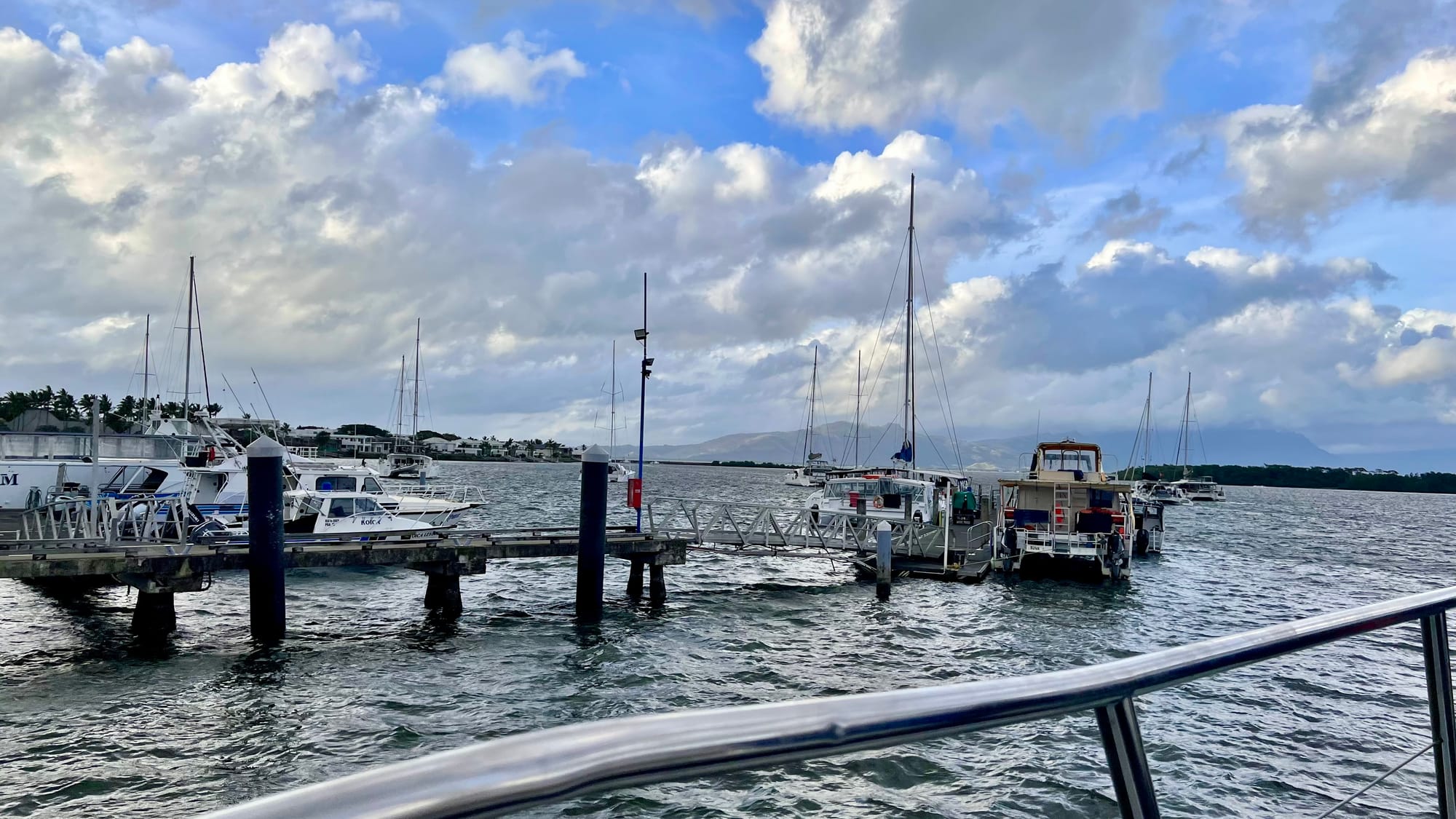 Port Denarau marina seen over a boat’s railing, with wooden jetties, moored motorboats and sailboat masts under a sky full of clouds.