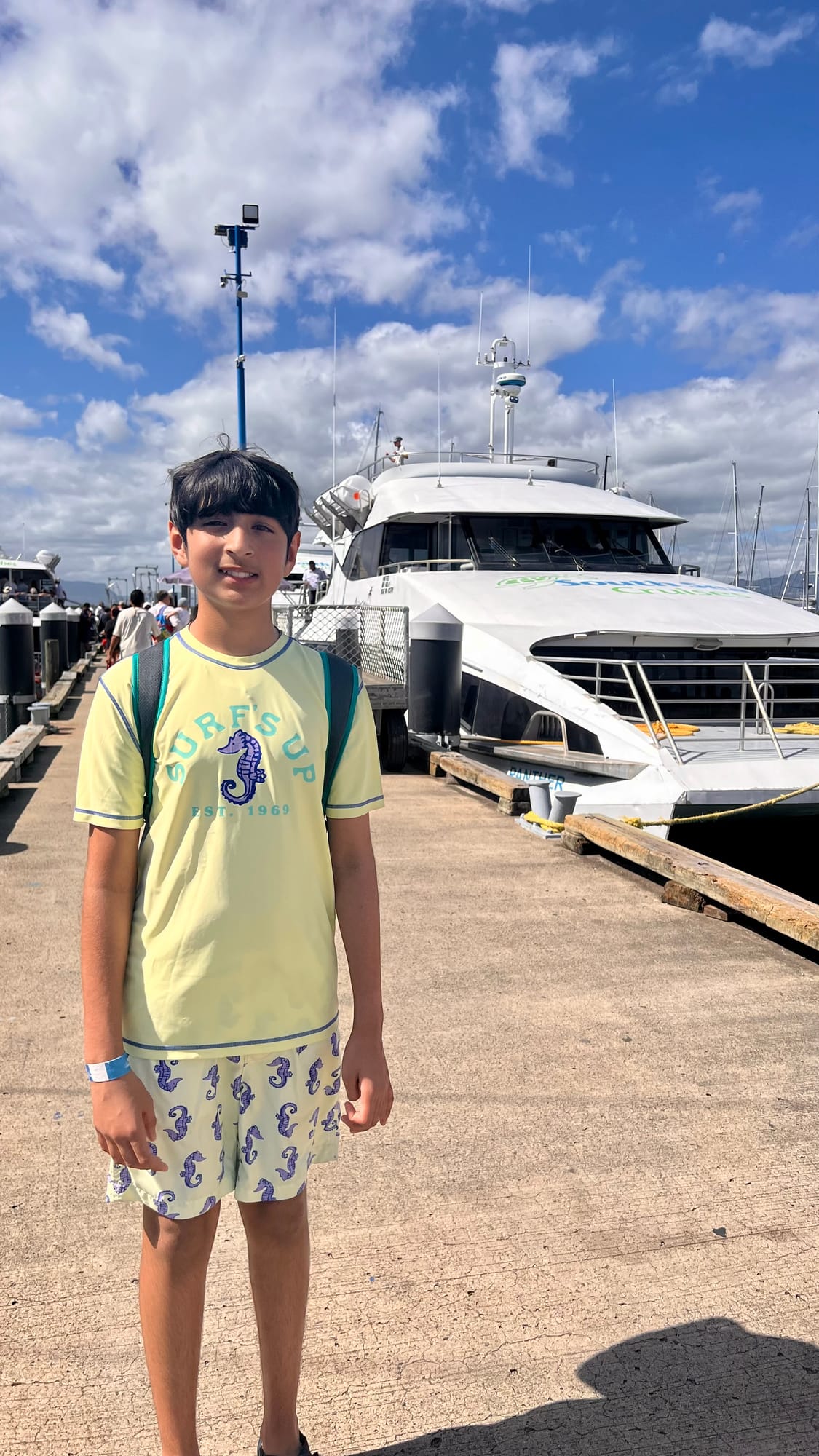Our son standing on the wharf in a yellow T-shirt and seahorse shorts, in front of a large white catamaran.