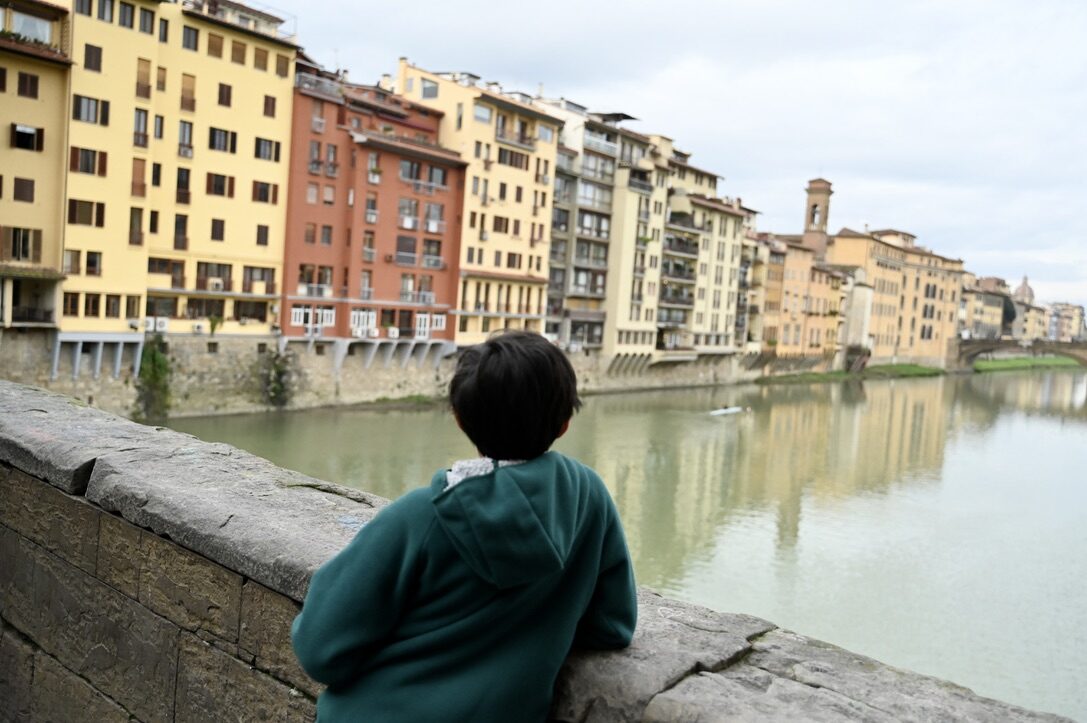 Boy leaning on the wall of the Ponte Vecchio, looking along the Arno at the colorful riverside houses
