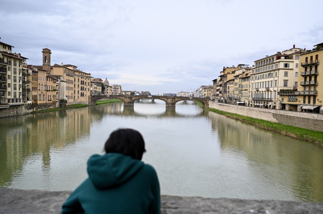 Boy on a bridge looking at a bridge over a river in the distance with tuscan buildings on both sides of the bank