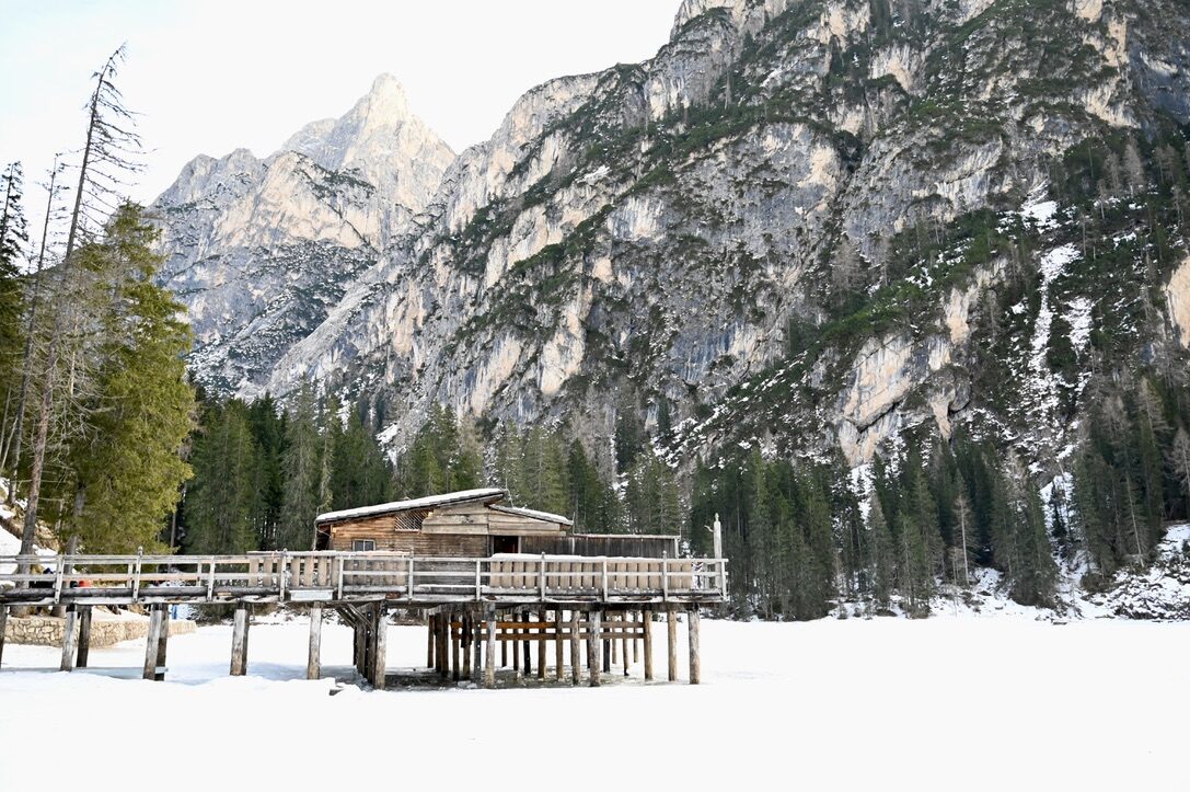 A wooden boathouse on the snow-covered, frozen lake below rocky peaks