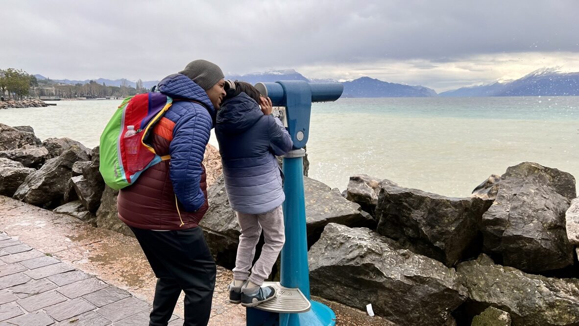 Woman and boy at a coin-operated viewer on the rocky lakeshore, with mountains across the water