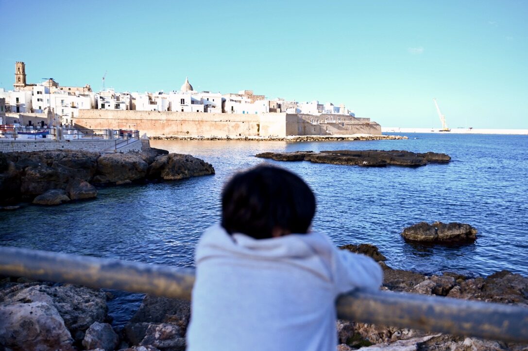 Boy at a railing looking across the water to Monopoli’s white old town