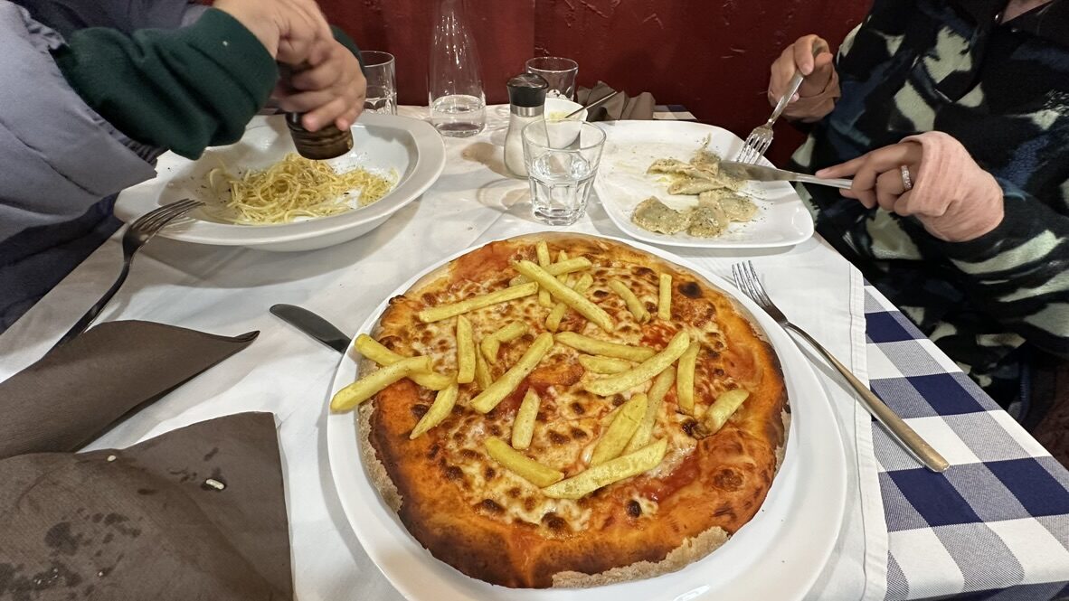 A plate of pizza topped with fries and a bowl of spaghetti on a checked tablecloth
