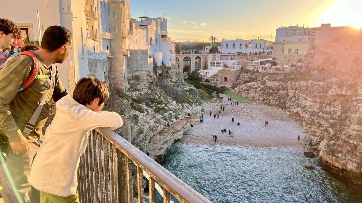 Man and boy at a balcony railing looking down at the beach between the cliffs at sunset