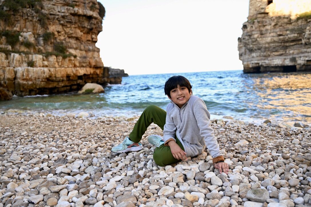 Boy sitting on the pebble beach below the tall cliffs of Polignano a Mare
