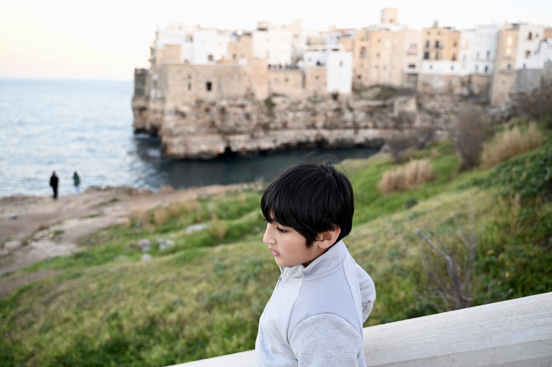 Boy on the side of a walkway with grass covered sea-side cliff on one side and the other side of the cliff is rugged with European style stone buildings on it