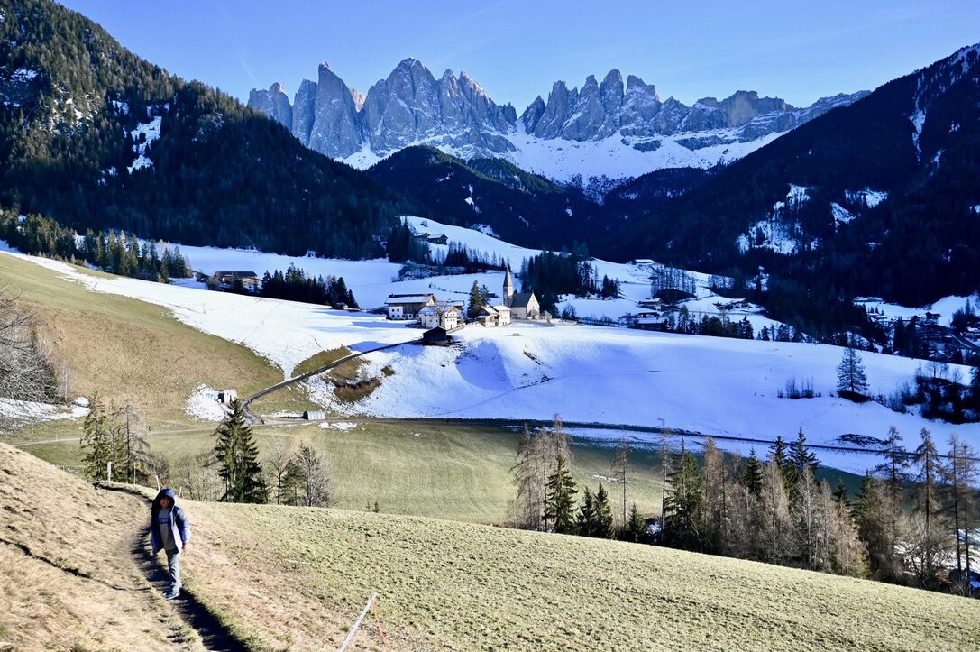 A hiker on a grassy path above the village and its church, with the jagged Odle peaks behind