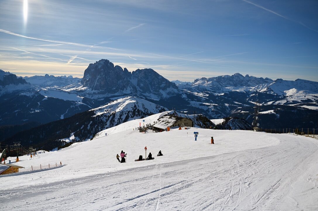 A wide ski slope with skiers, and a range of Dolomite peaks on the horizon