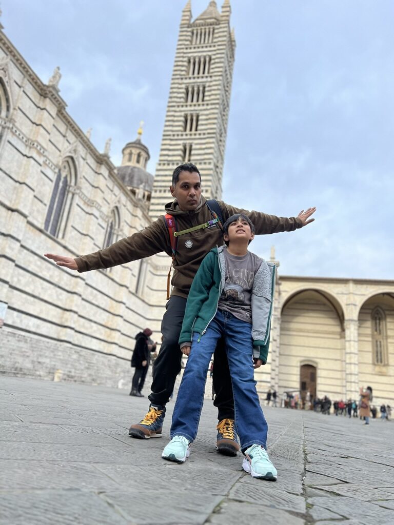 Man and boy with arms out in front of the striped bell tower of Siena Cathedral