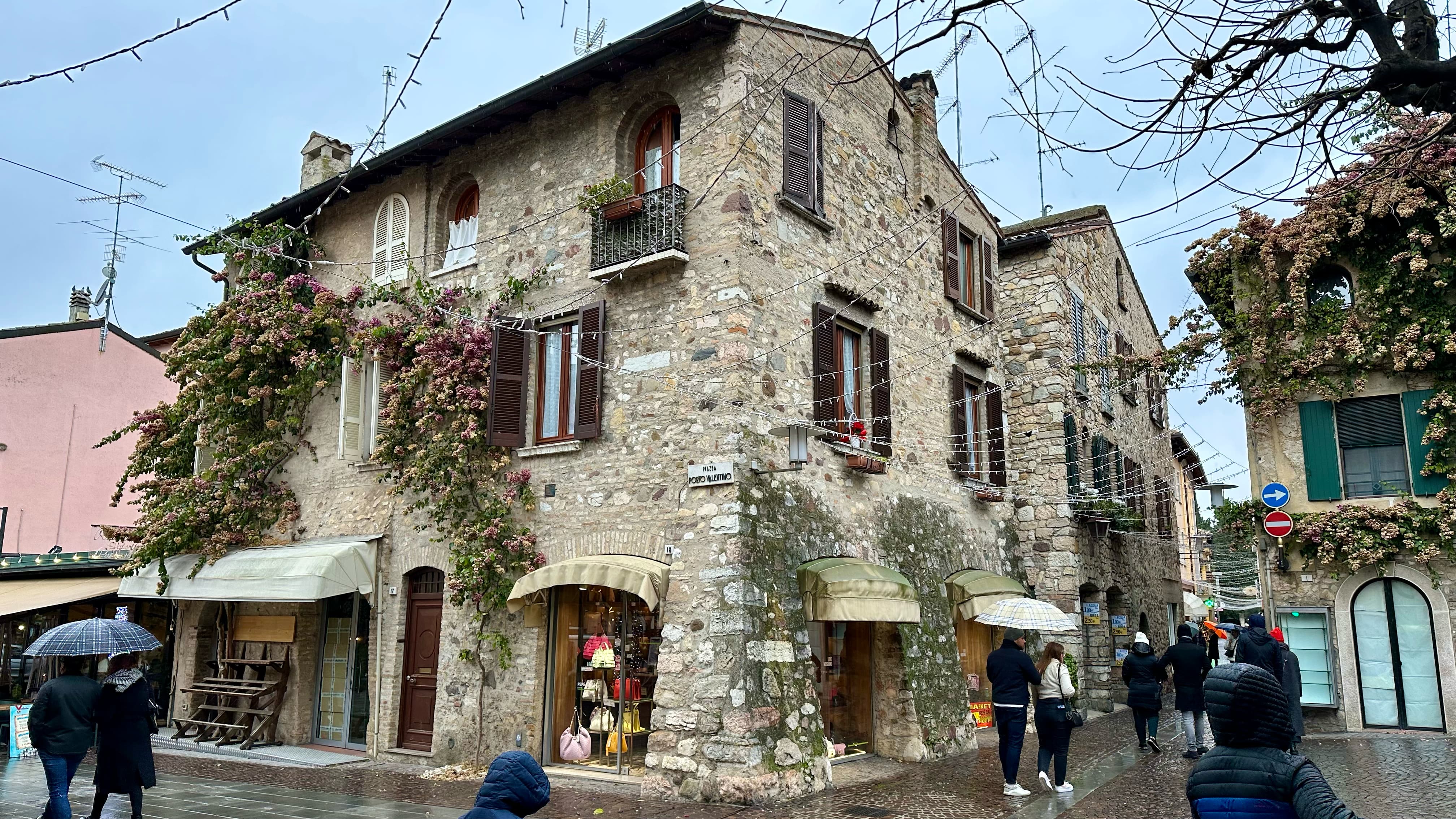 European style stone building with shops on the bottom level and creeper bushes with flowers covering some of the walls, with people walking around the cobble stone pathways