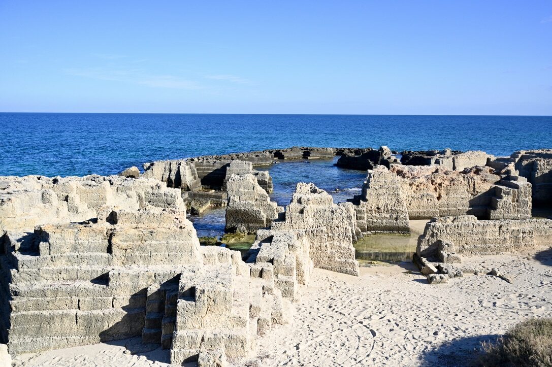 Pale limestone ruins cut into the rocks beside the blue sea
