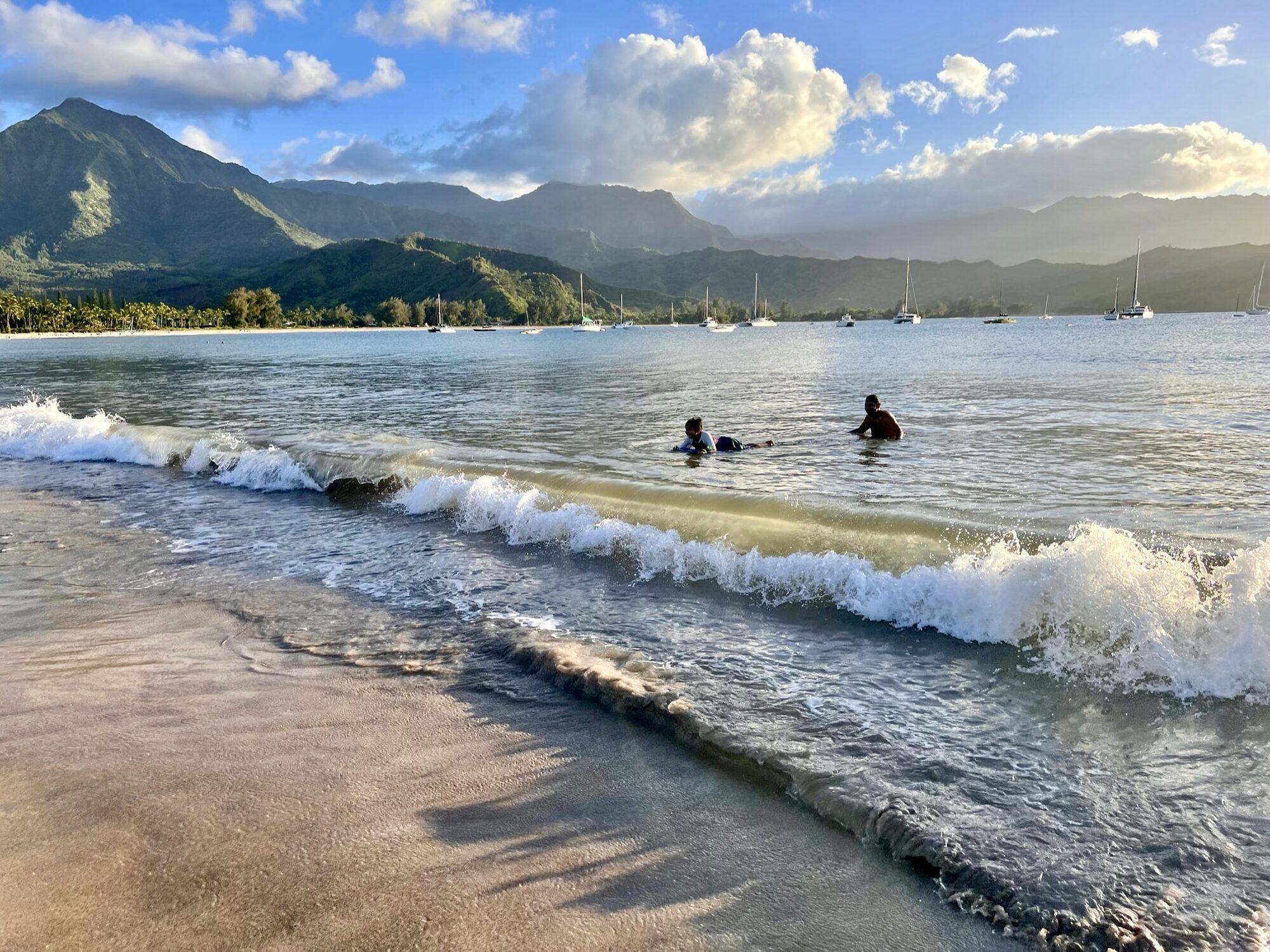 Swimmers in Hanalei Bay with sailboats moored below green mountains