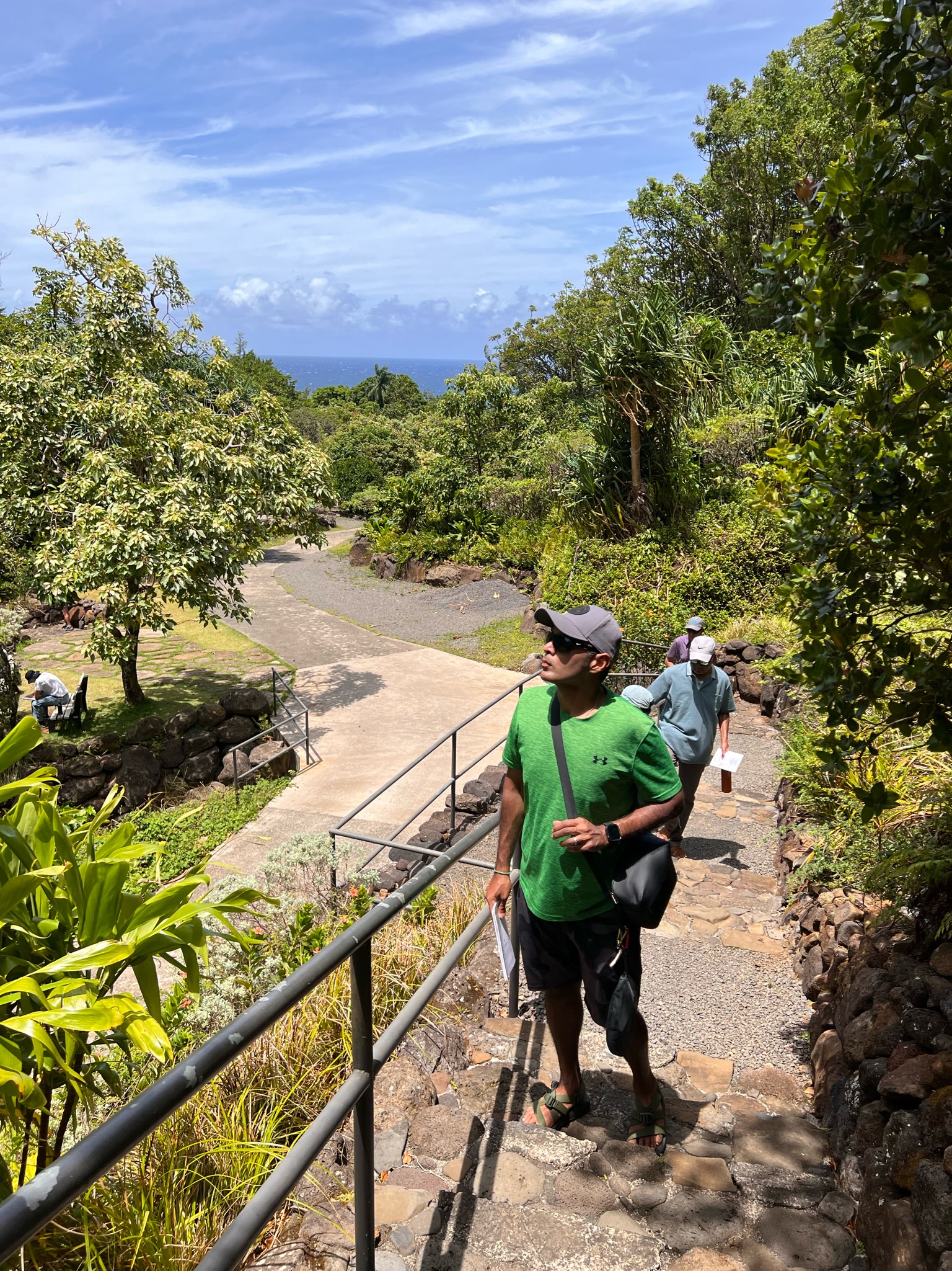 _Man in green shirt on a walkway inside a tropical garden with family members in the background and ocean in the distance.