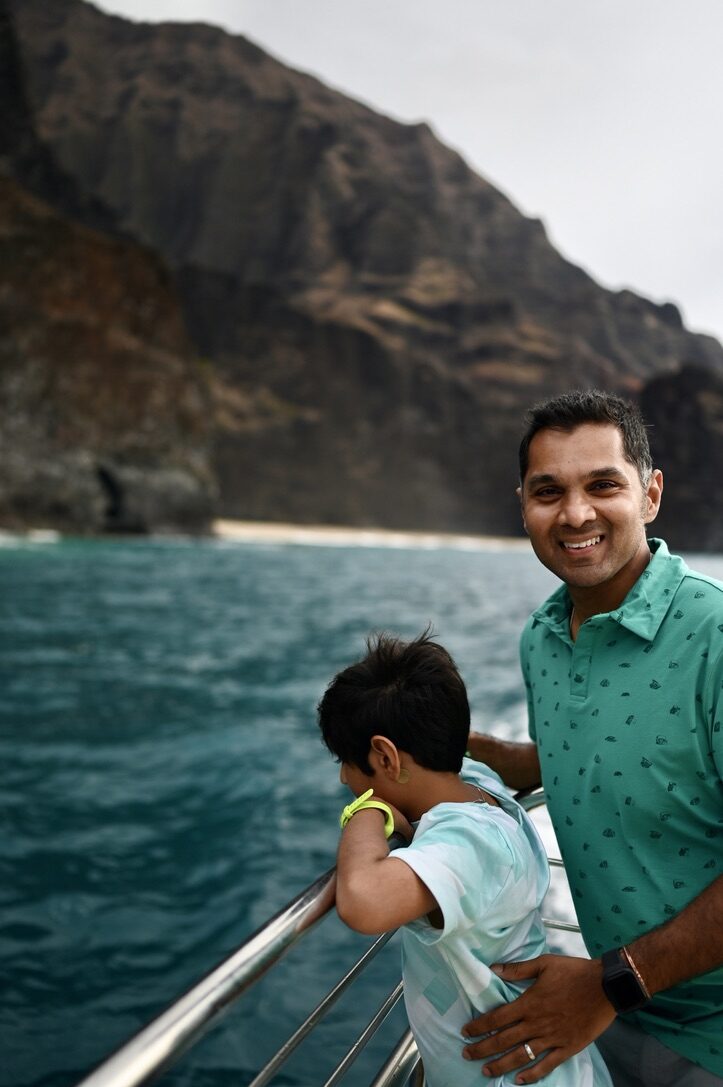 Man and boy at the boat railing beside the dark cliffs of the Napali Coast