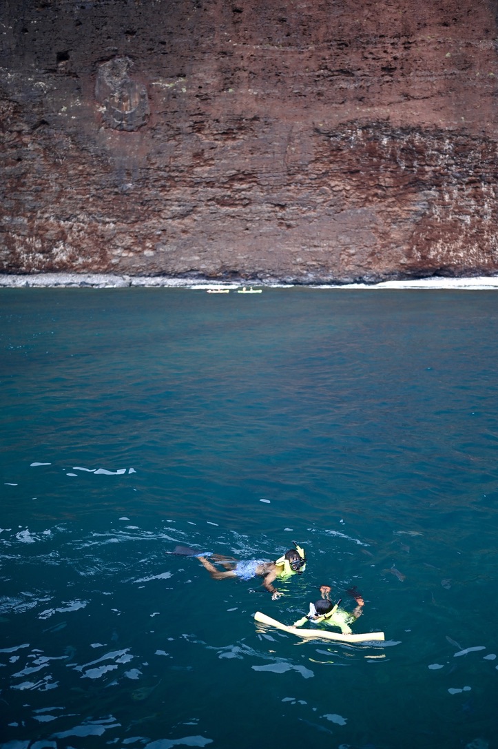 Boy and man snorkeling in beautiful dark turquoise blue waters along sheer red cliffs and a narrow beach