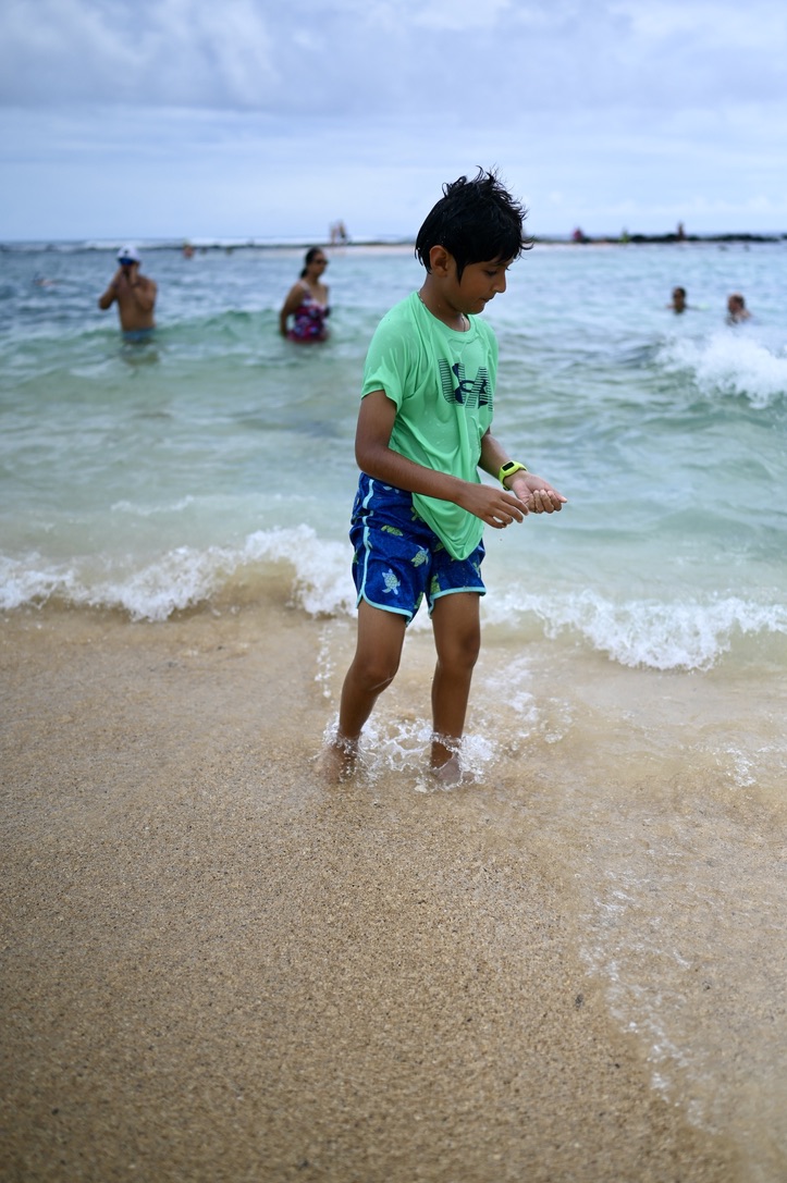 Boy standing on sandy beach with turquoise beach in the background with people in the water