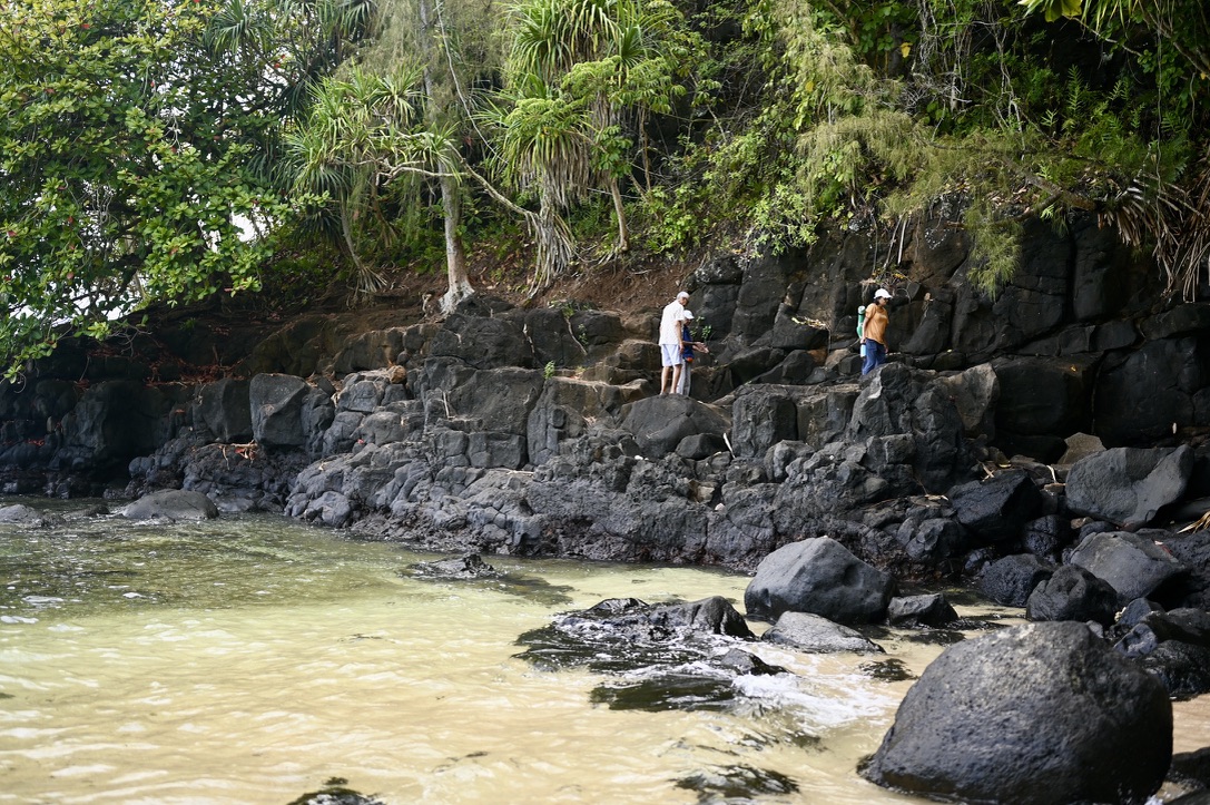 _2 elderly men and an elder woman along a rocky hill side with big boulders, by a bay of the ocean.