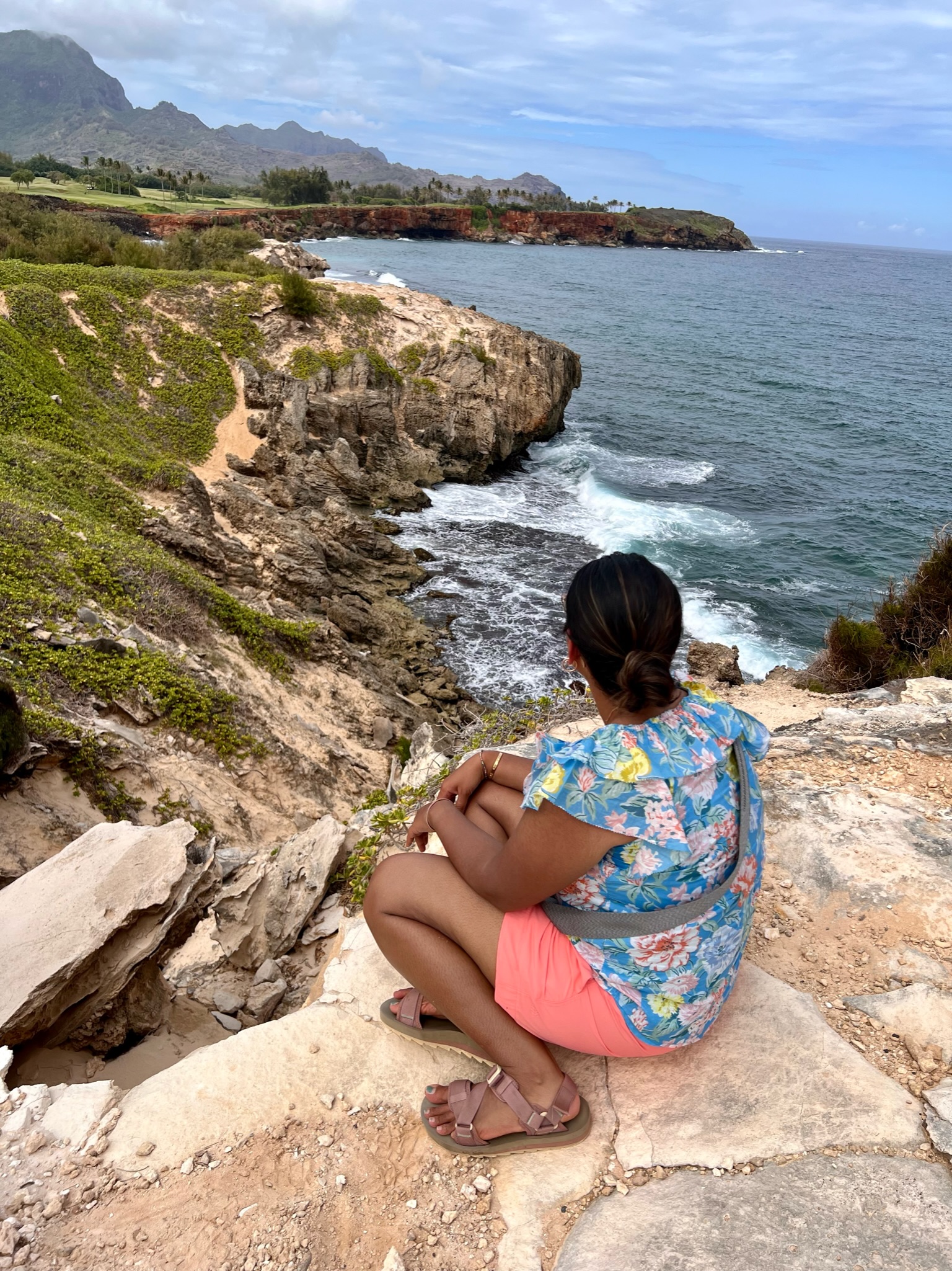 Woman sitting on a rock ledge with her back to the camera looking at a rugged coastline with golf course and mountains in the distance; brown sand and ground cover plants cover the path heading into the distance